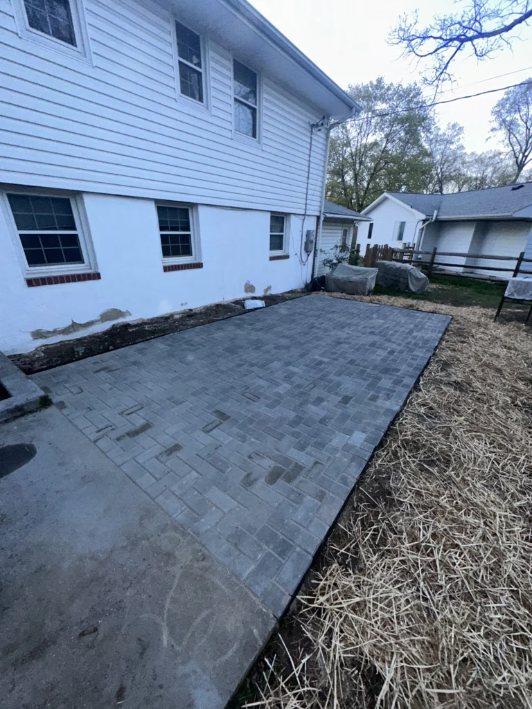 Newly installed gray paver patio alongside a white house with straw covering surrounding soil